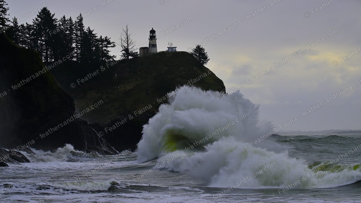 A large wave bursting against a headland below a cliff-top lighthouse.
